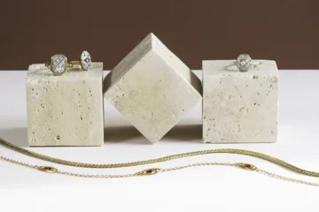 Trois cubes en marbre blanc sur lesquels sont posés trois bagues. Devant deux colliers en maille dorés sont posés en S sur un fond blanc