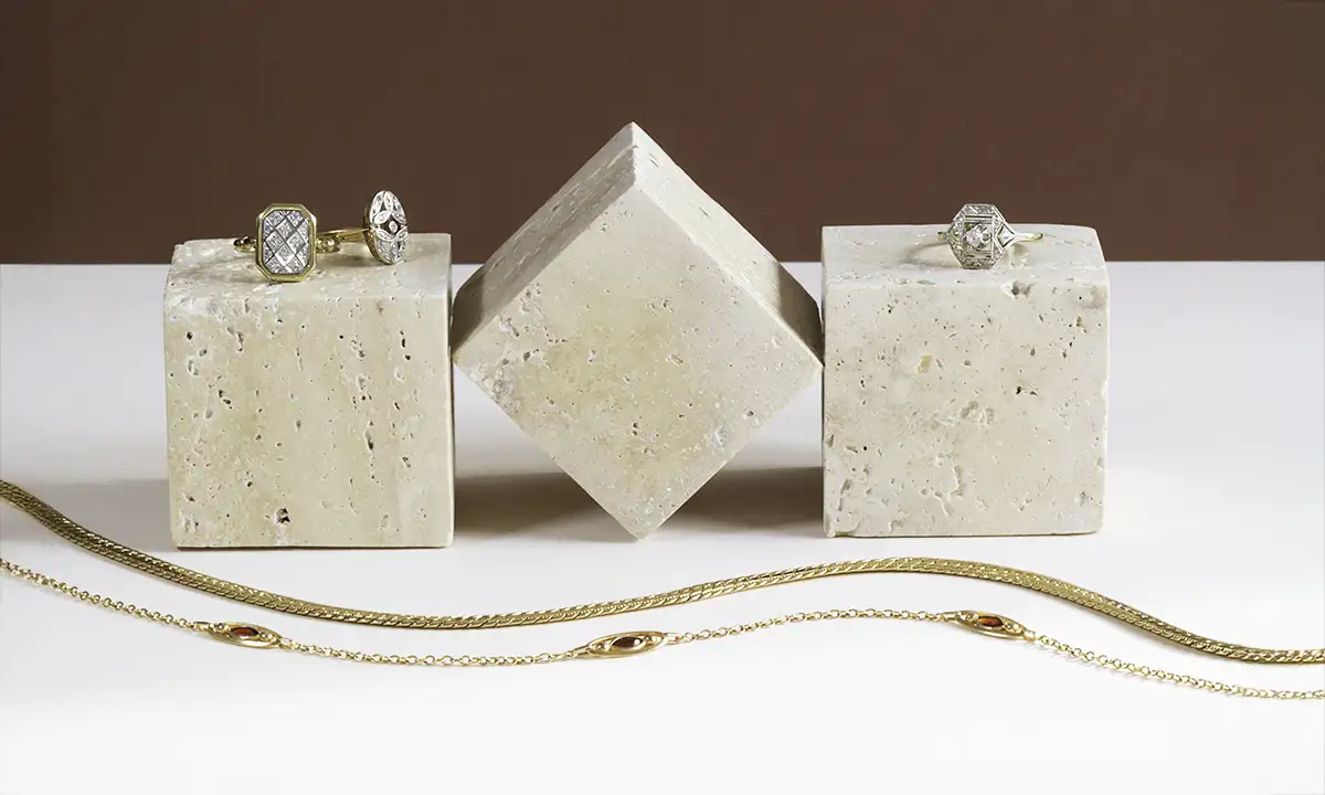 Trois cubes en marbre blanc sur lesquels sont posés trois bagues. Devant deux colliers en maille dorés sont posés en S sur un fond blanc
