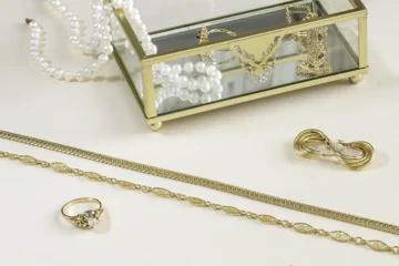 Boite en métal dorée et verre ouverte contenant des bijoux anciens en or et un collier de perles, devant des bijoux sont posés sur un fond beige