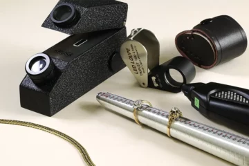 Plusieurs outils de gemmologie déposés sur un fond blanc neutre. Refractomètre, loupe, triboulet.