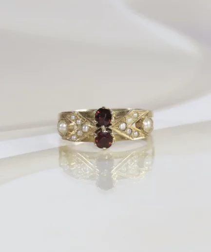 Bague ancienne en or rose surmontée de deux grenats ronds entourés de perles fines. La bague est posée sur un fond neutre beige