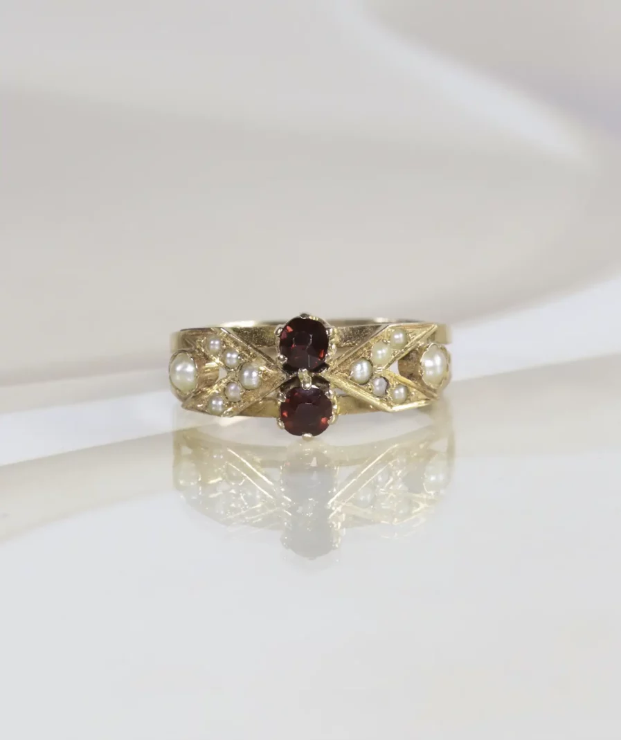 Bague ancienne en or rose surmontée de deux grenats ronds entourés de perles fines. La bague est posée sur un fond neutre beige