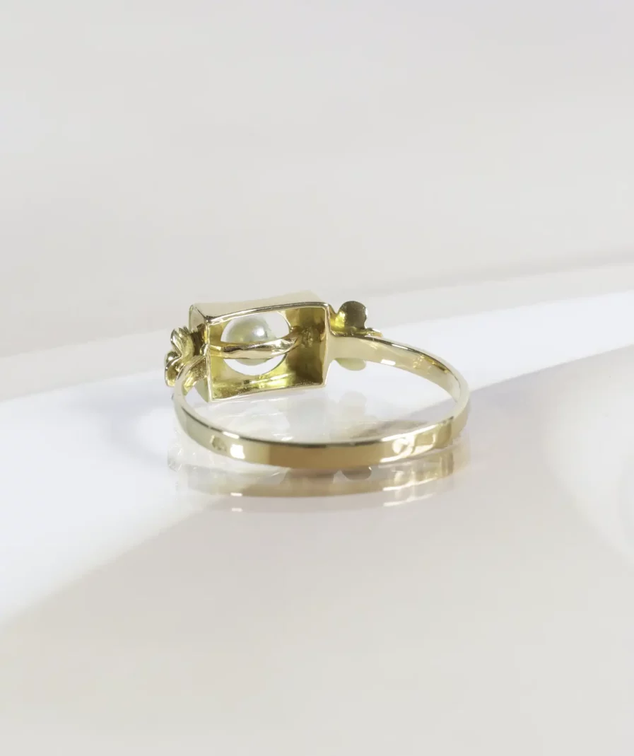 Bague vintage en or, motif rectangulaire avec une perle de culture en son centre, de part et d'autre une fleur en or. La bague est posée de dos sur un fond neutre beige.