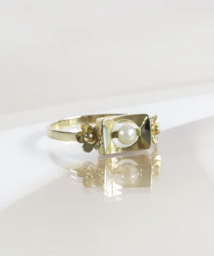 Bague vintage en or, motif rectangulaire avec une perle de culture en son centre, de part et d'autre une fleur en or. La bague est posée légèrement de profil sur un fond neutre beige.