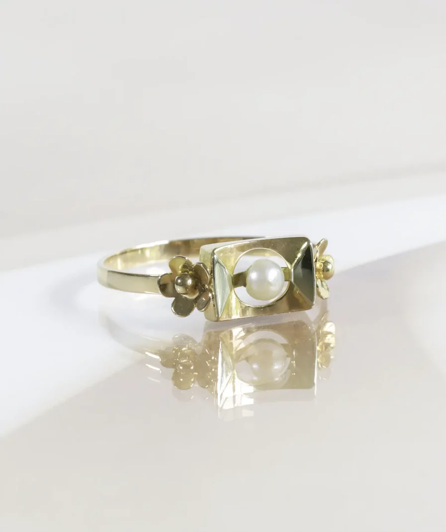Bague vintage en or, motif rectangulaire avec une perle de culture en son centre, de part et d'autre une fleur en or. La bague est posée légèrement de profil sur un fond neutre beige.