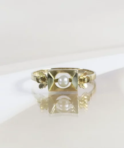 Bague vintage en or, motif rectangulaire avec une perle de culture en son centre, de part et d'autre une fleur en or. La bague est posée sur un fond neutre beige.