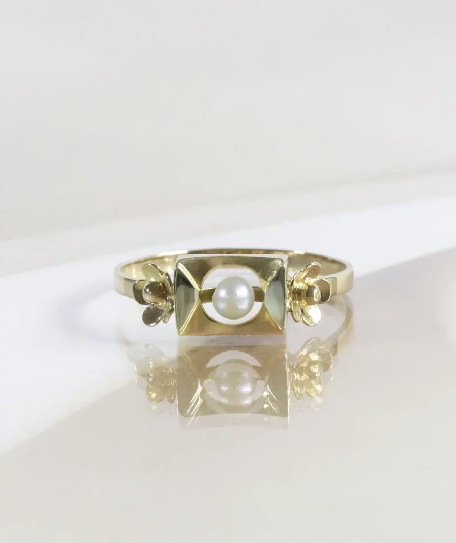 Bague vintage en or, motif rectangulaire avec une perle de culture en son centre, de part et d'autre une fleur en or. La bague est posée sur un fond neutre beige.