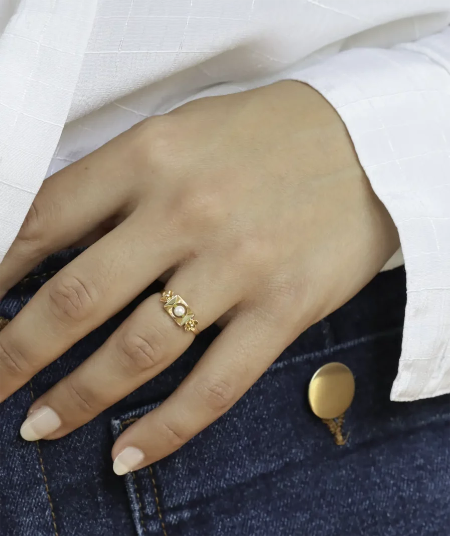Bague en or deux tons or et blanc. De forme rectangulaire avec une perle de culture en son centre. La bague est disposée sur l'annulaire d'une main qui repose sur un jean.